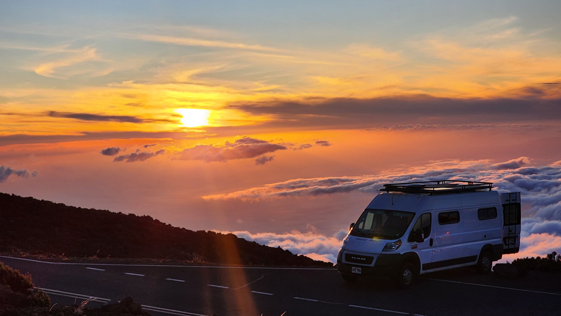 A white camper van parked on a mountain road above a sea of clouds during a bright, golden sunset.