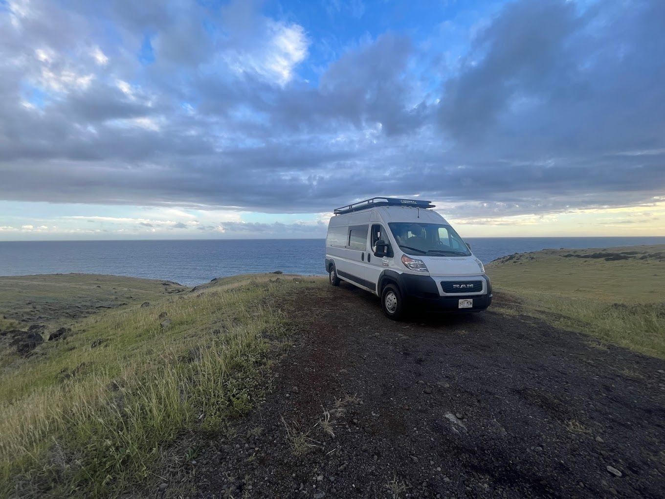 A white camper van parked on a grassy, volcanic coastal cliff overlooking the ocean under a cloudy sky.