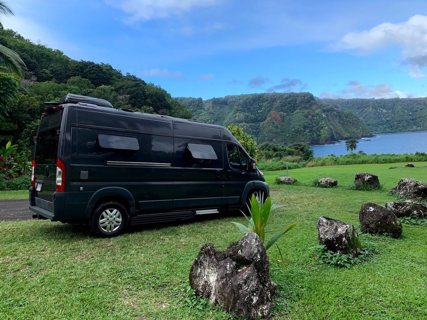 A dark-colored camper van parked on a grassy field overlooking a bay, with rocky outcroppings and mountains in the distance.