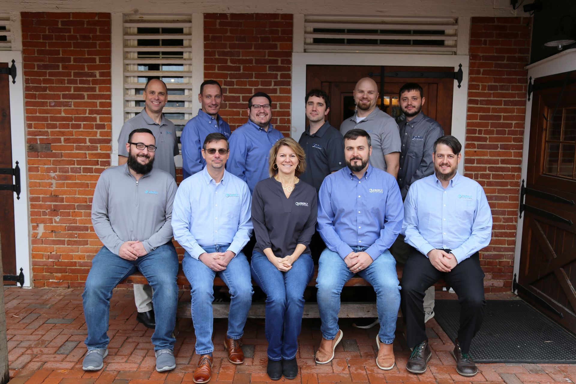 Group of eleven people posed on a wooden bench in front of a brick building.