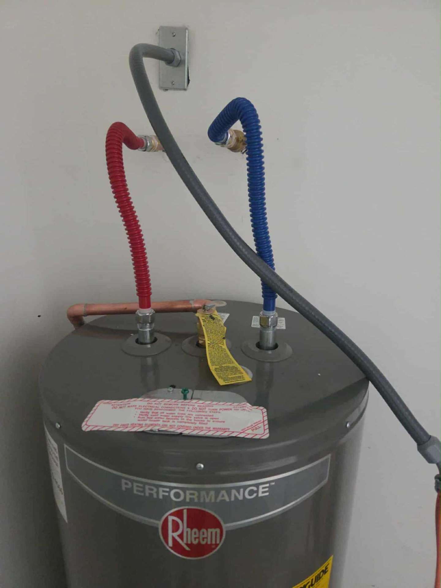 A Rheem water heater with red and blue flexible water lines, installed against a white wall in a garage.