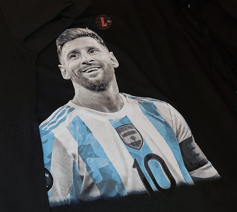 Una persona con una medalla y la camiseta argentina sostiene dos trofeos. Fondo negro.