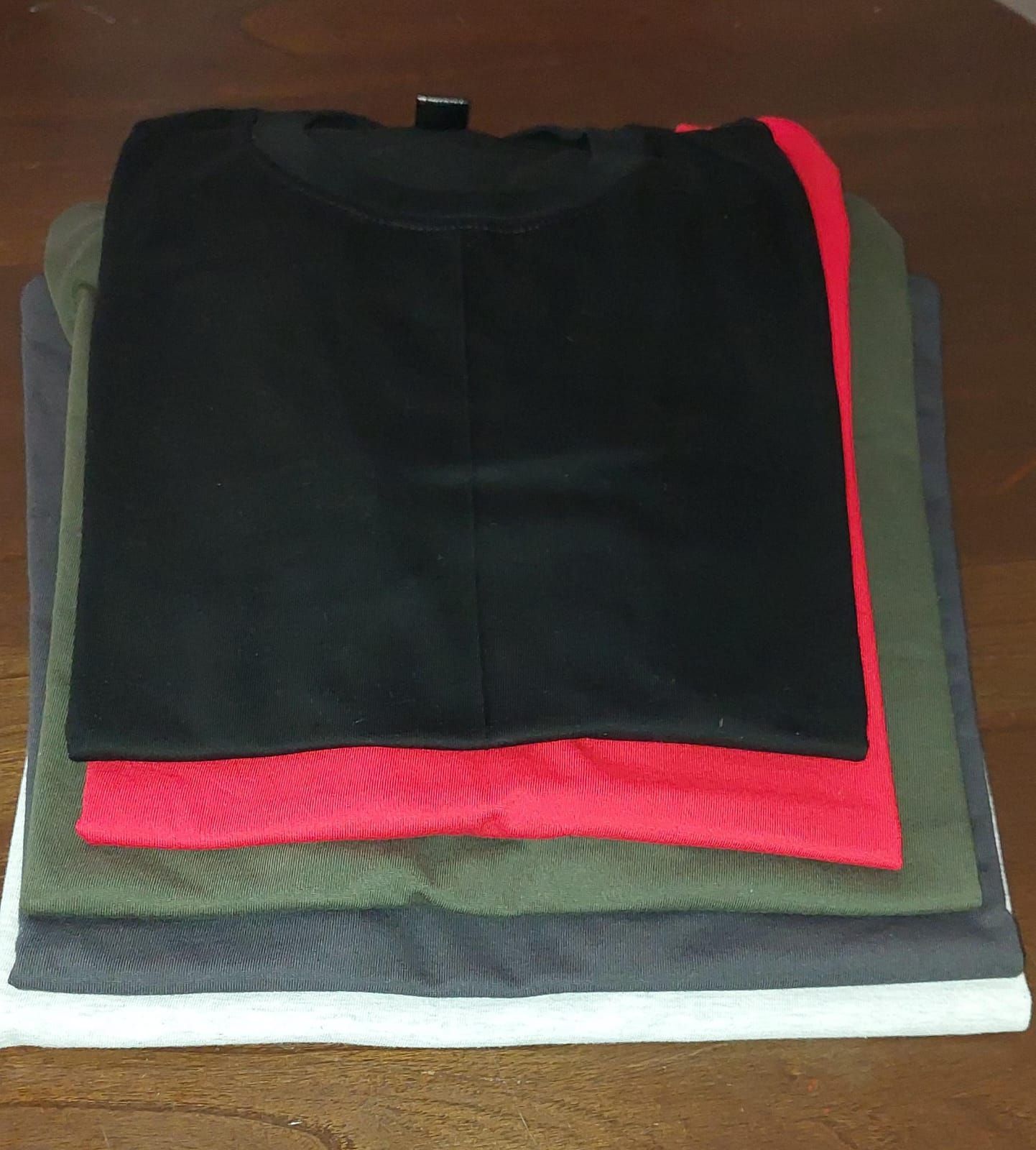 Camisetas apiladas y dobladas en varios colores: negro, rojo, verde, gris y blanco, sobre una superficie de madera.