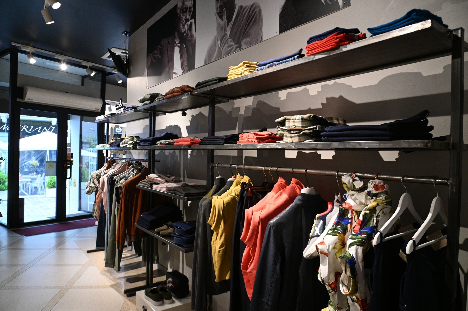 Interno di un negozio di abbigliamento con appendiabiti e scaffali per esporre i capi. Pareti scure, porte in vetro e illuminazione a incasso.