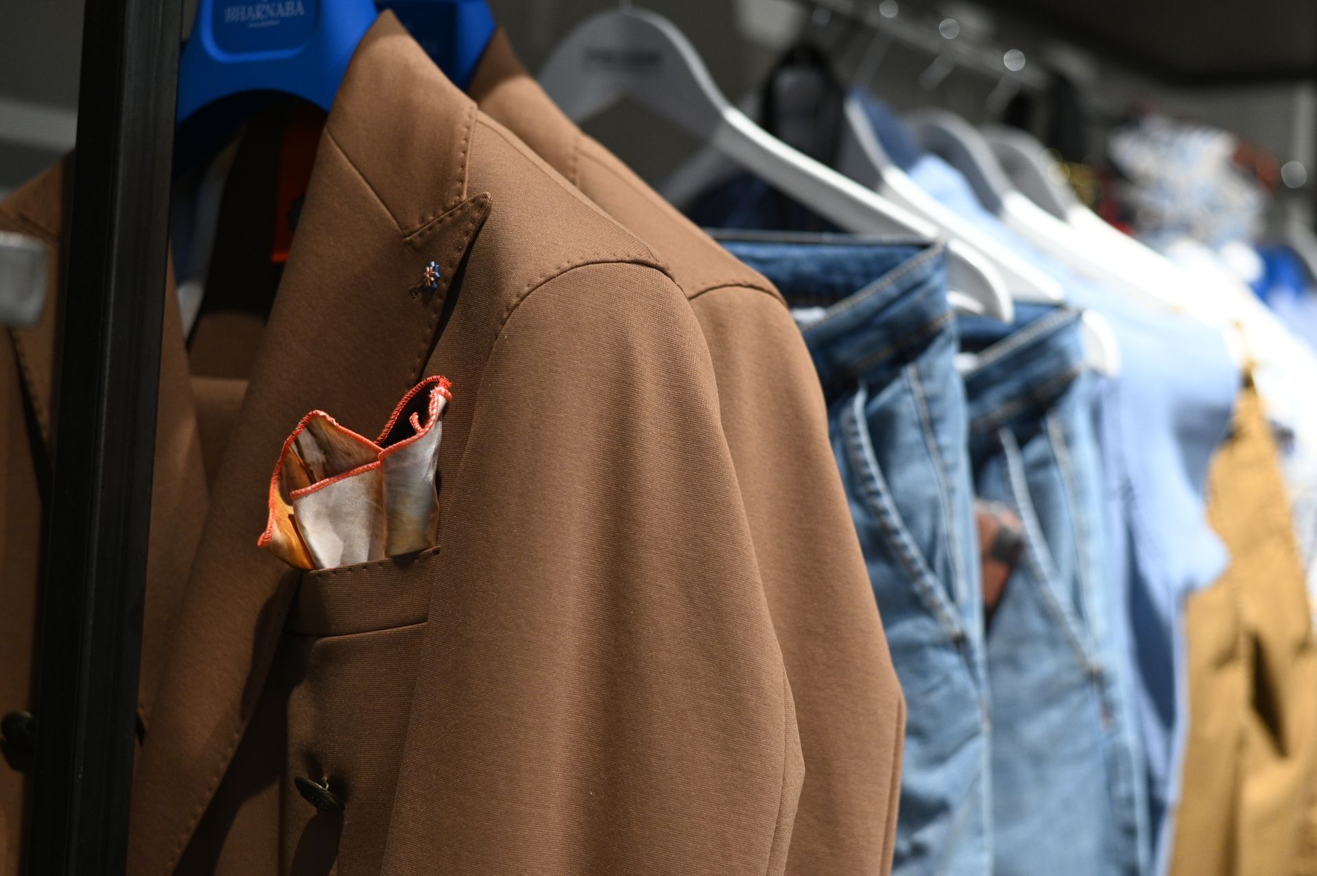 Un blazer marrone con fazzoletto da taschino è appeso su un attaccapanni insieme a jeans e altri indumenti in un negozio.