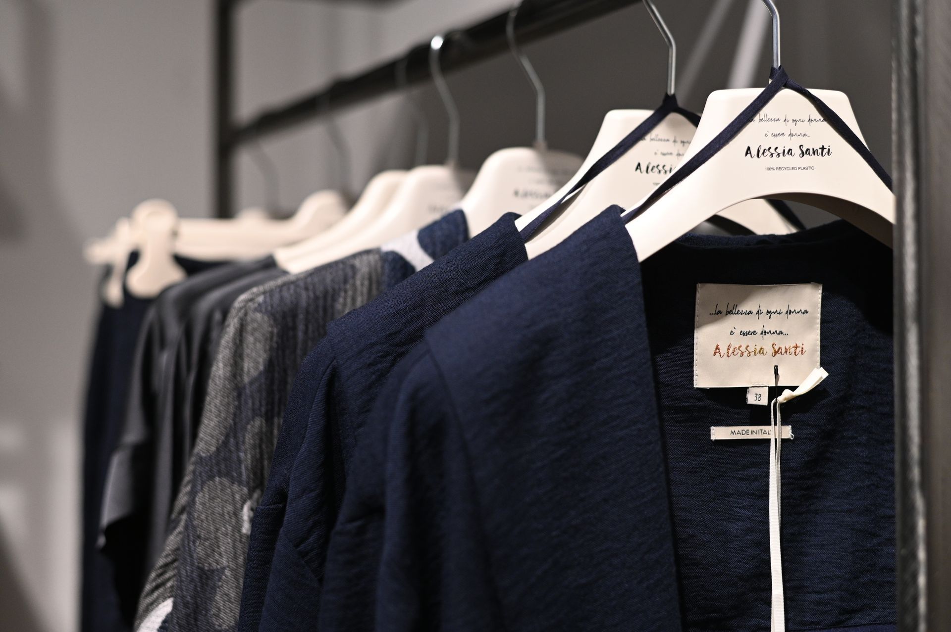 Abiti appesi su un attaccapanni in una boutique. Capi blu navy, grigi e neri su grucce bianche.