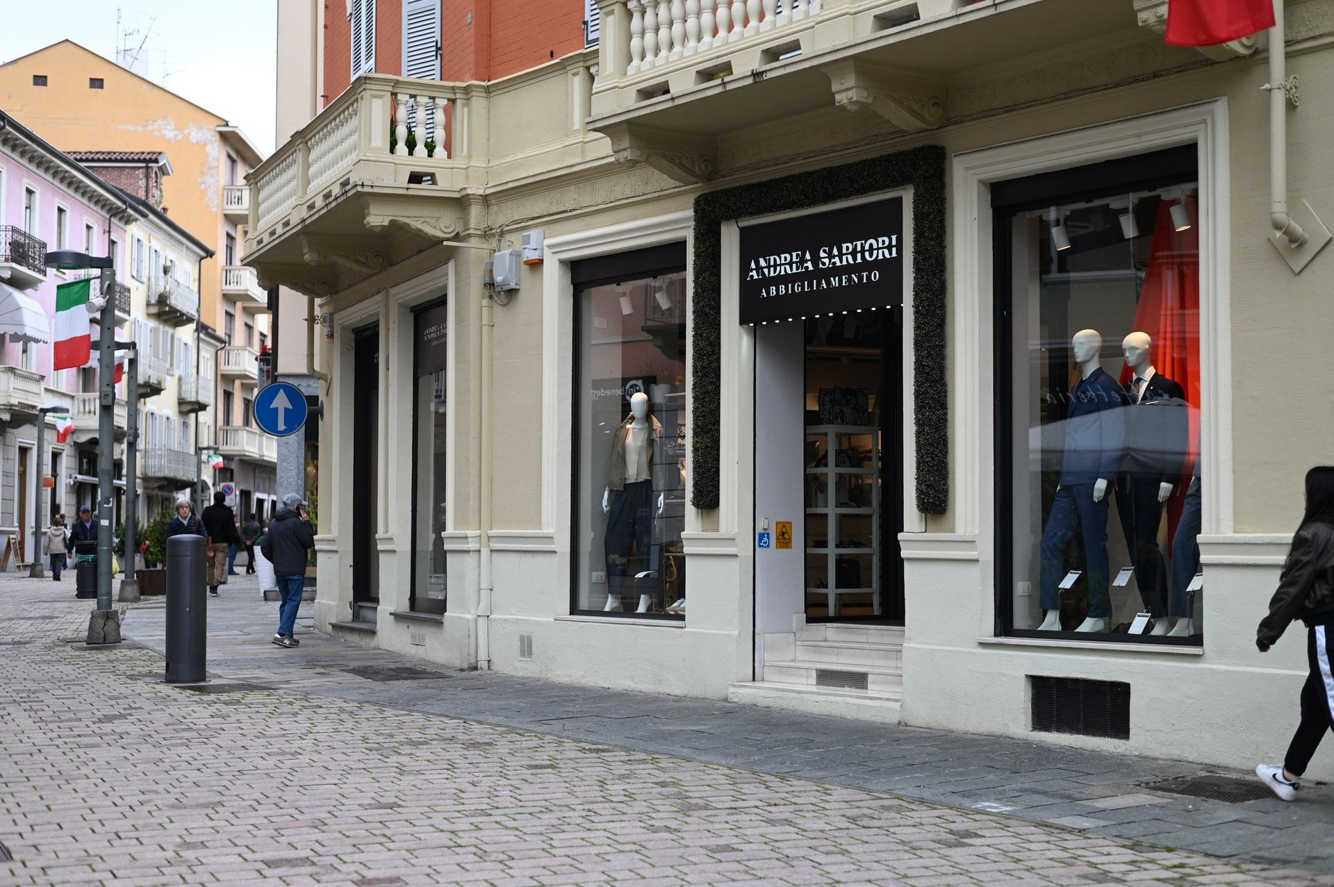 Scena di strada con negozio di abbigliamento; manichini in vetrina, persone che camminano sui ciottoli.