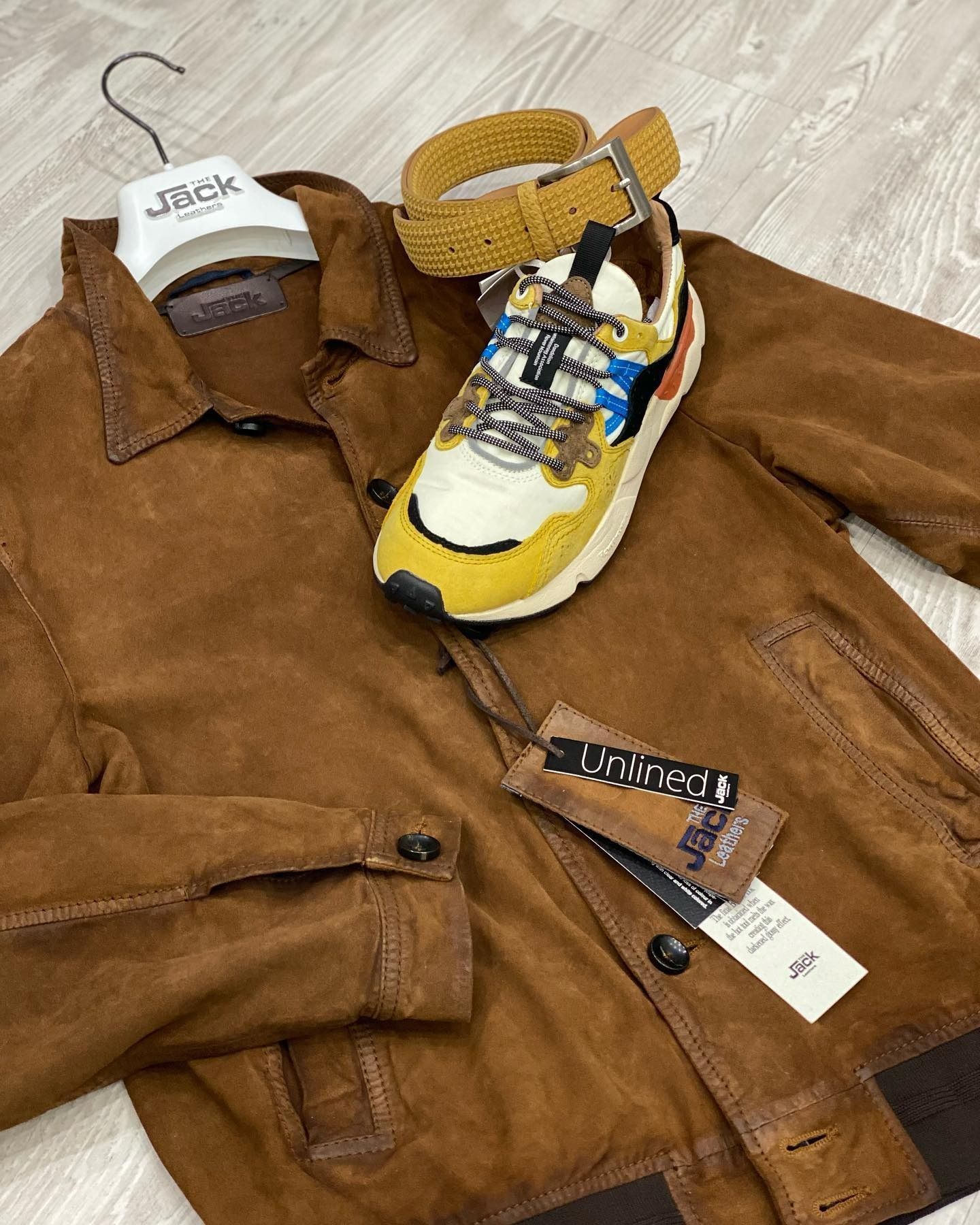 Giacca, sneaker e cintura in pelle scamosciata marrone esposte su una superficie di legno chiaro.