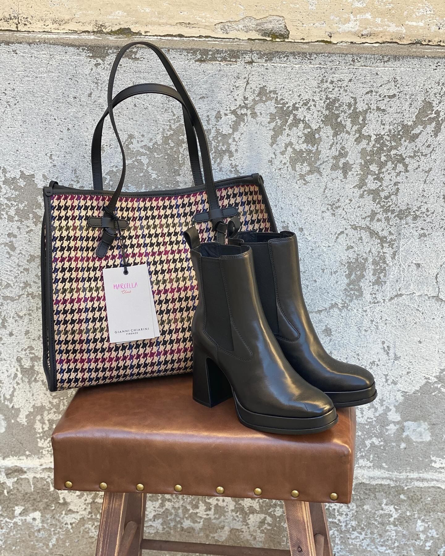 Borsa tote con motivo rosa, nero e beige, stivaletti neri e uno sgabello contro una parete ruvida.