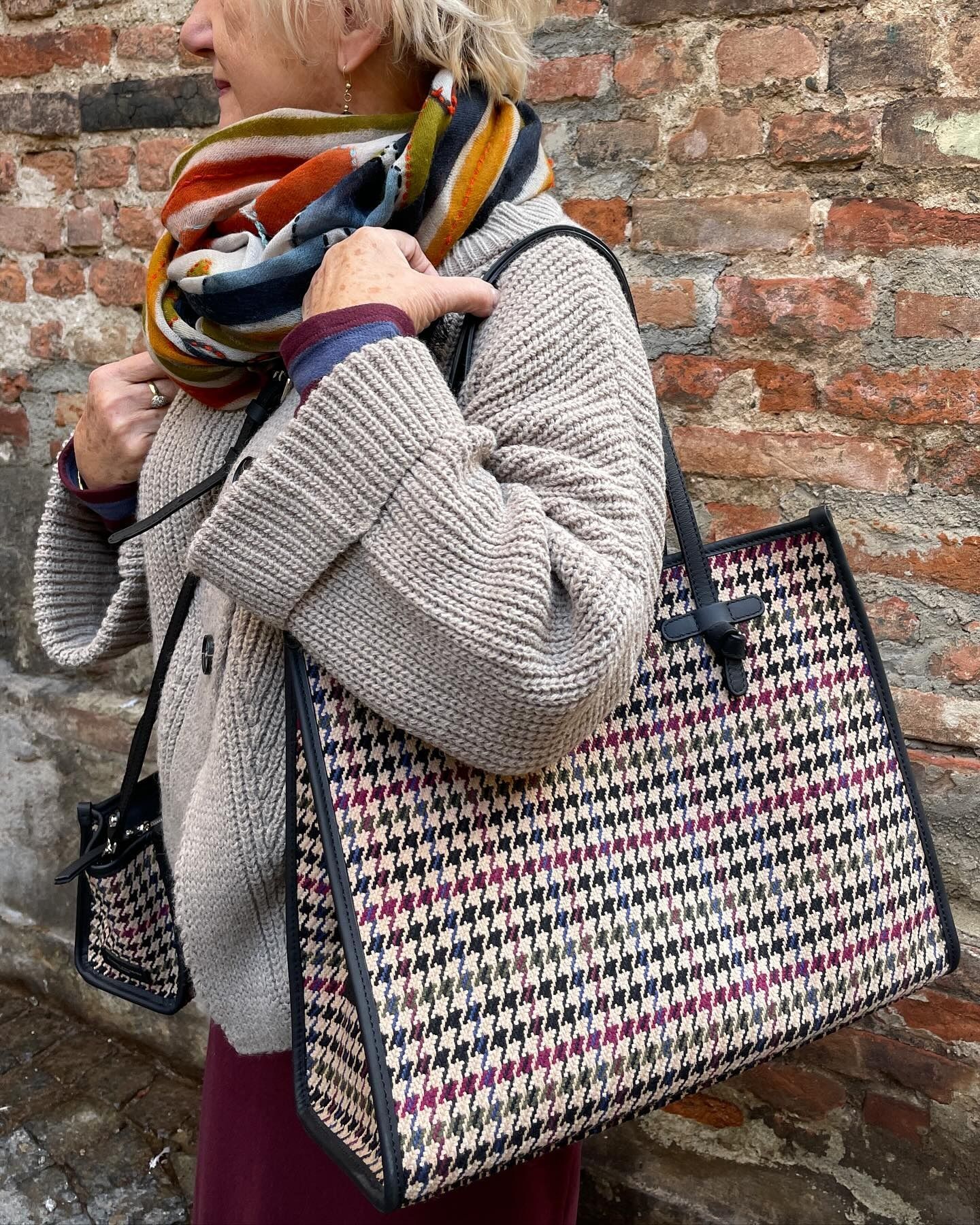 Donna che indossa una sciarpa e un maglione, con una grande borsa a tracolla fantasia e una piccola borsa coordinata, sfondo di mattoni.