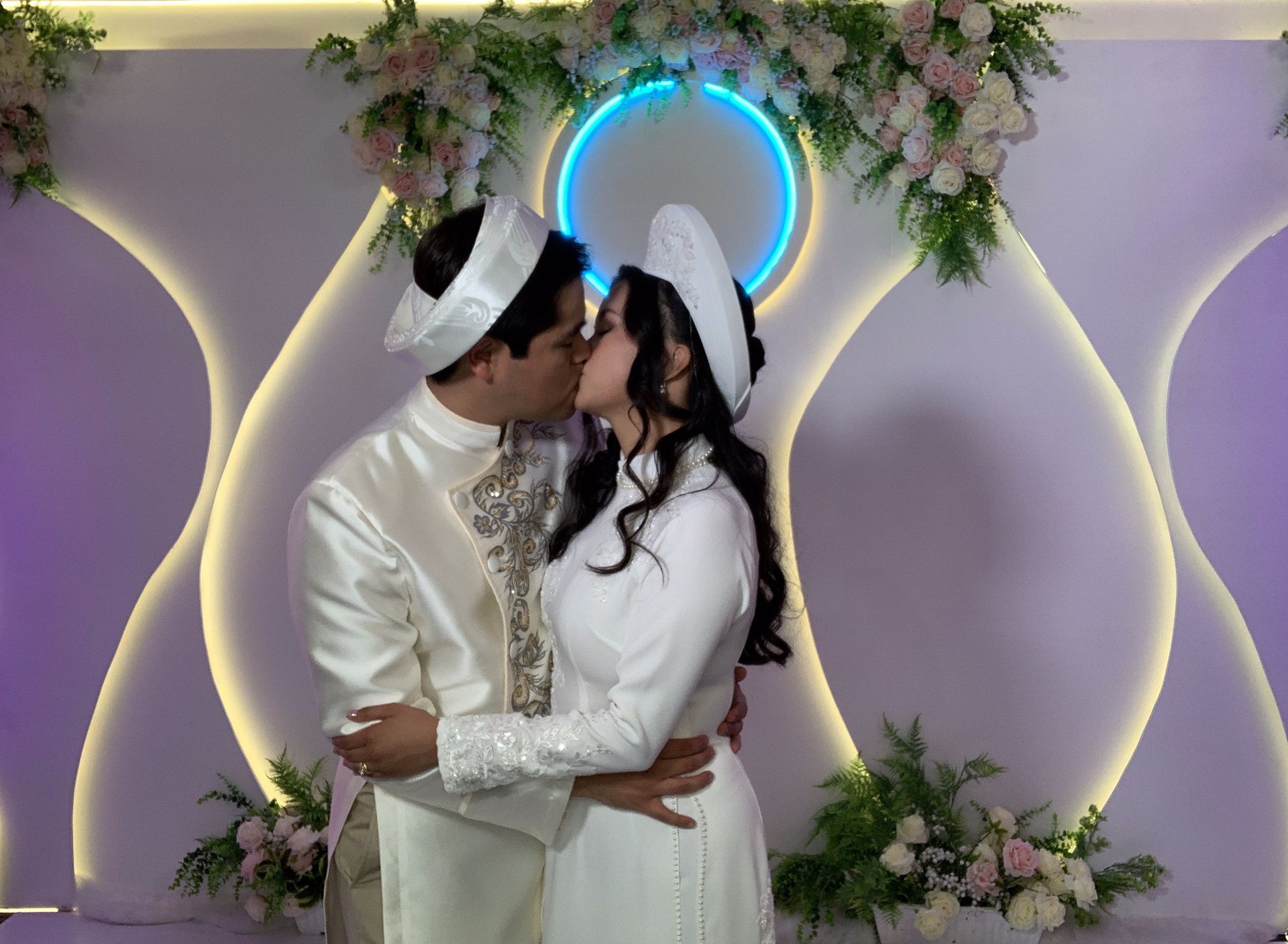 A couple in traditional white Vietnamese wedding attire kissing in front of a decorative, illuminated floral backdrop.
