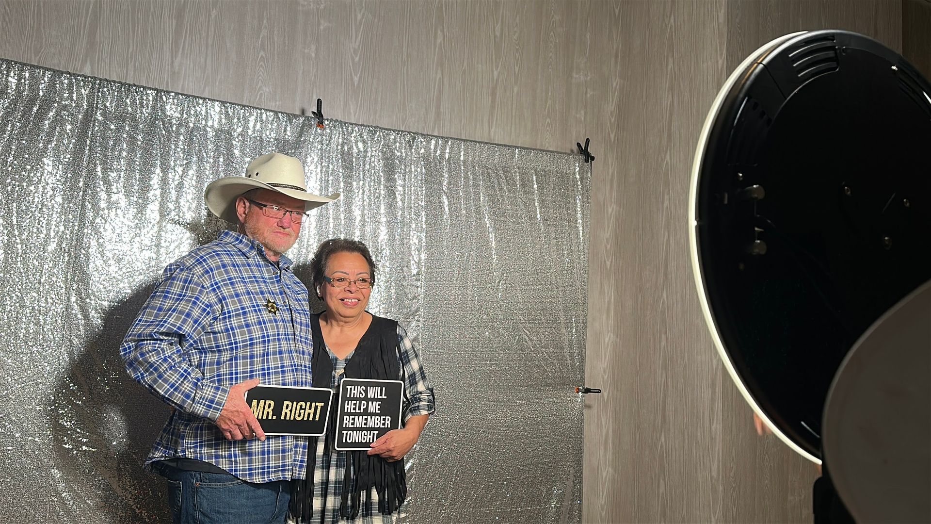 Couple in photo booth holding signs. Silver backdrop, large light, cowboy hat.