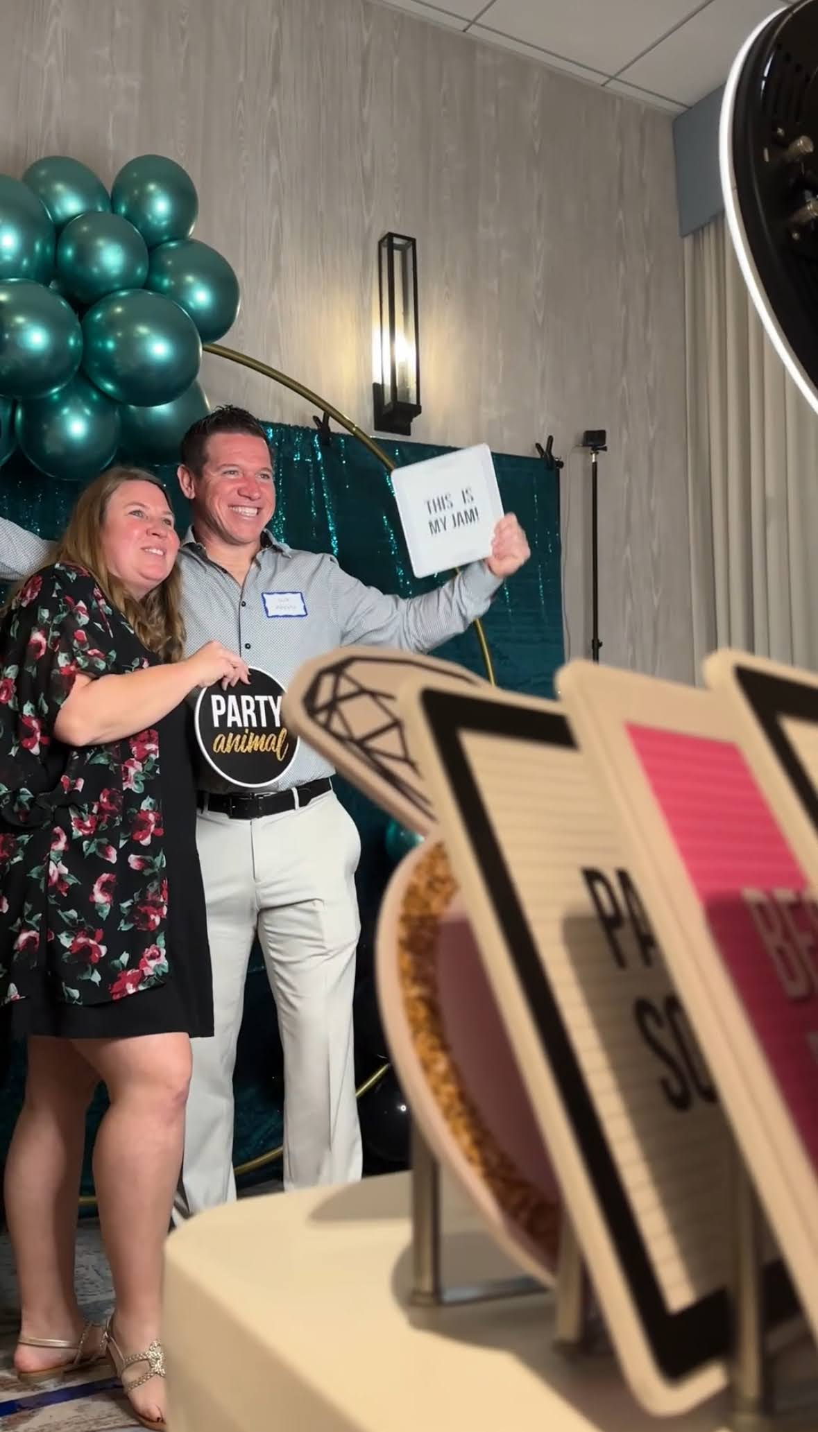 Woman and man posing at photo booth, holding props. Teal balloons and green backdrop.