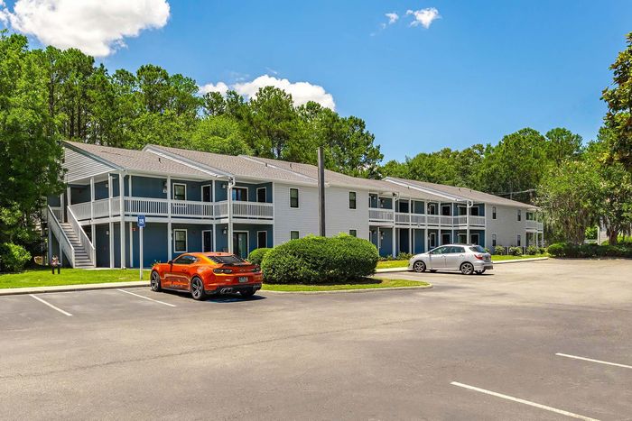 Two-story apartment building with cars parked in the lot, trees in the background, and blue siding.