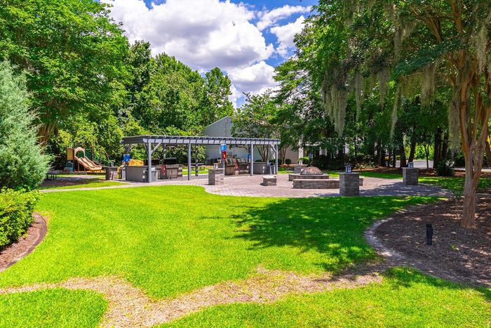 Lush green park with a playground, barbecue area, and trees under a partly cloudy sky.