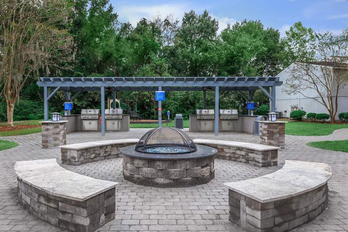 Outdoor grilling area with a fire pit surrounded by benches, under a pergola.