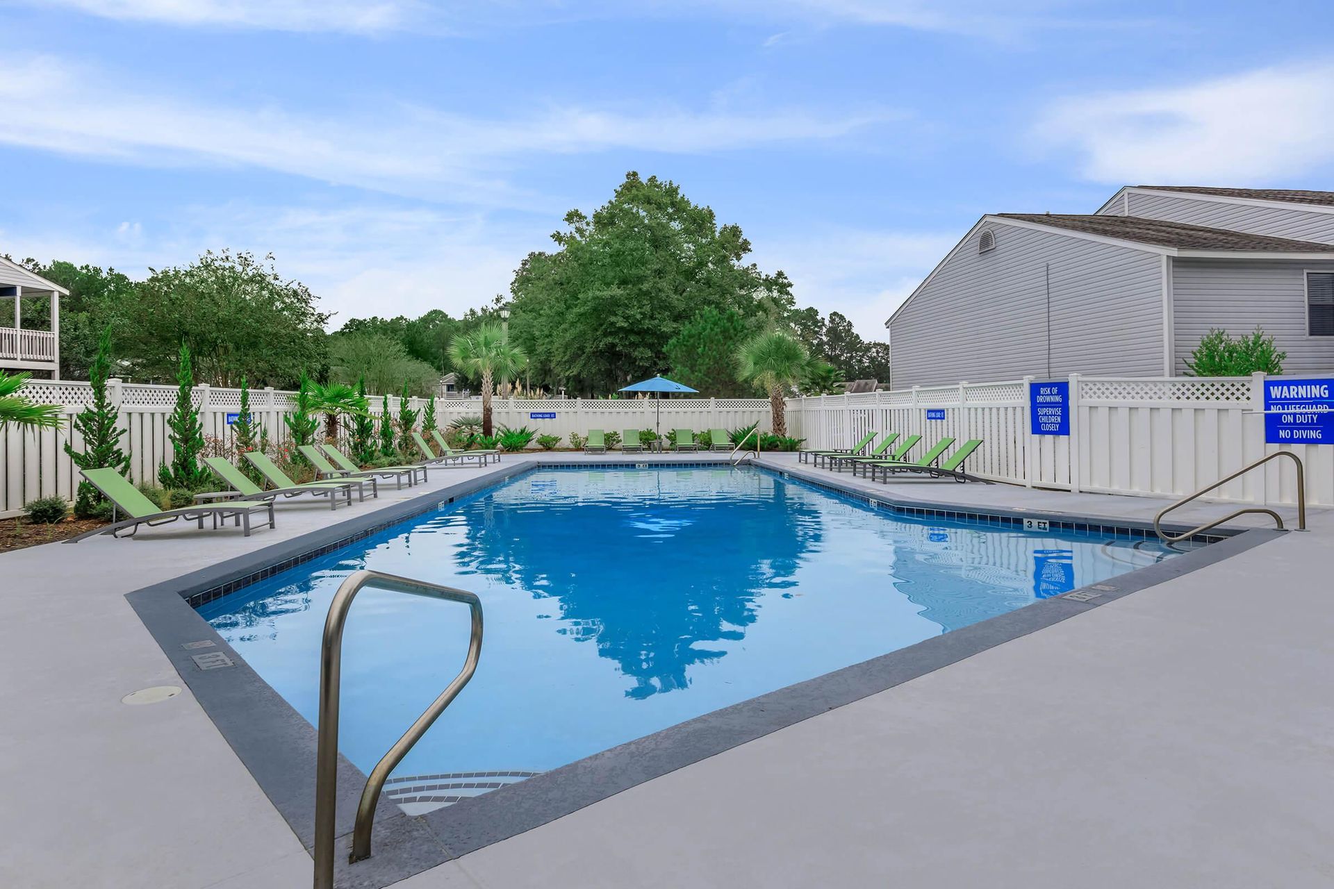 Pool area with lounge chairs, blue water, and white fence against a blue sky.