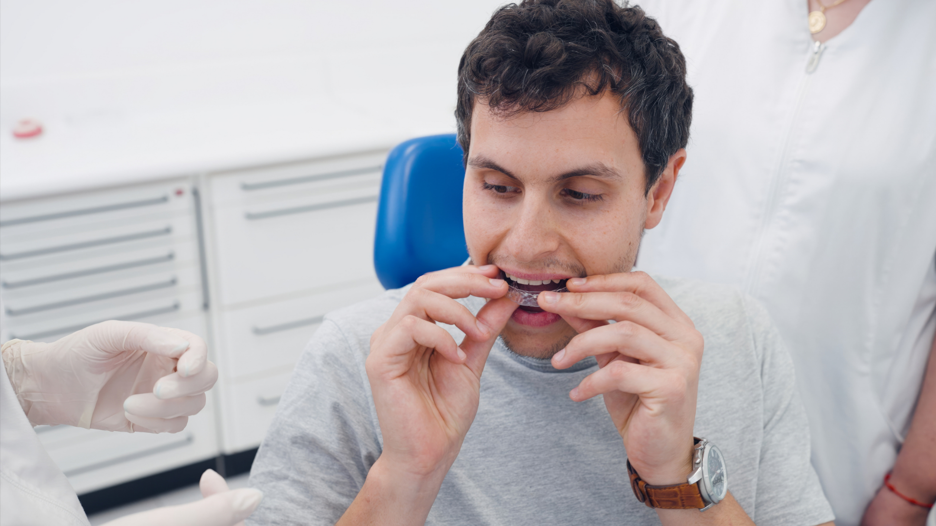 A patient sitting in a dental chair adjusting a clear aligner tray onto their teeth.