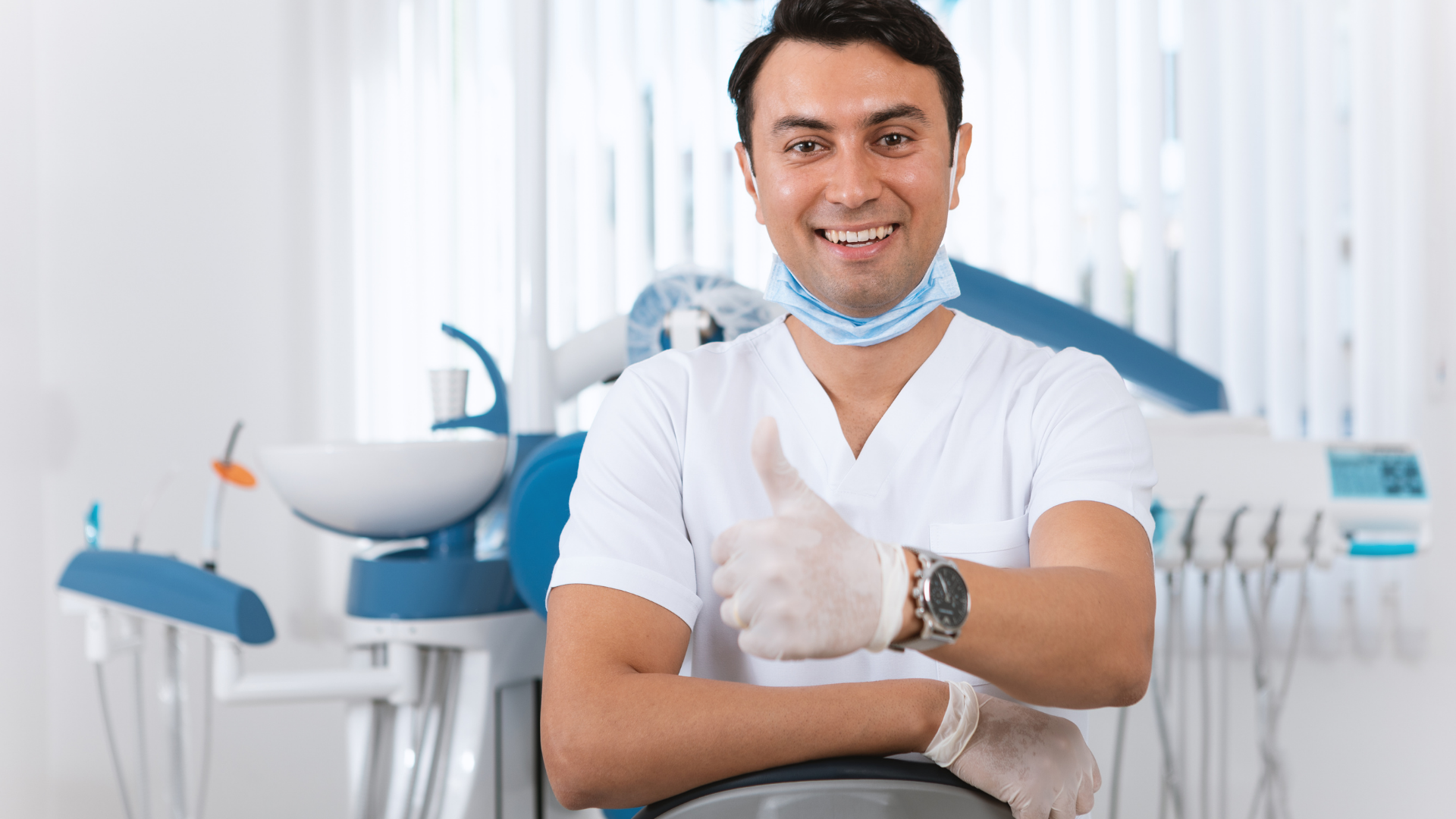Dentist in white scrubs gives thumbs up, smiling in a dental office with blue equipment.