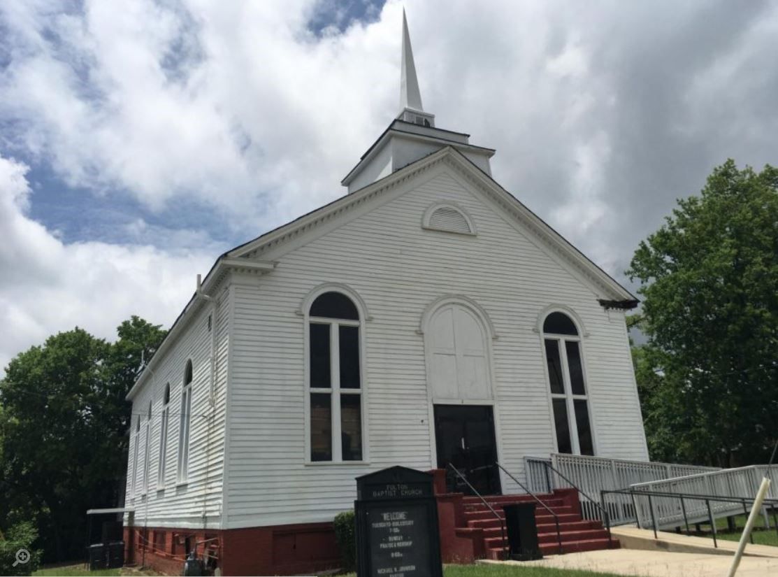 A small white church with a steeple is surrounded by trees on a cloudy day.