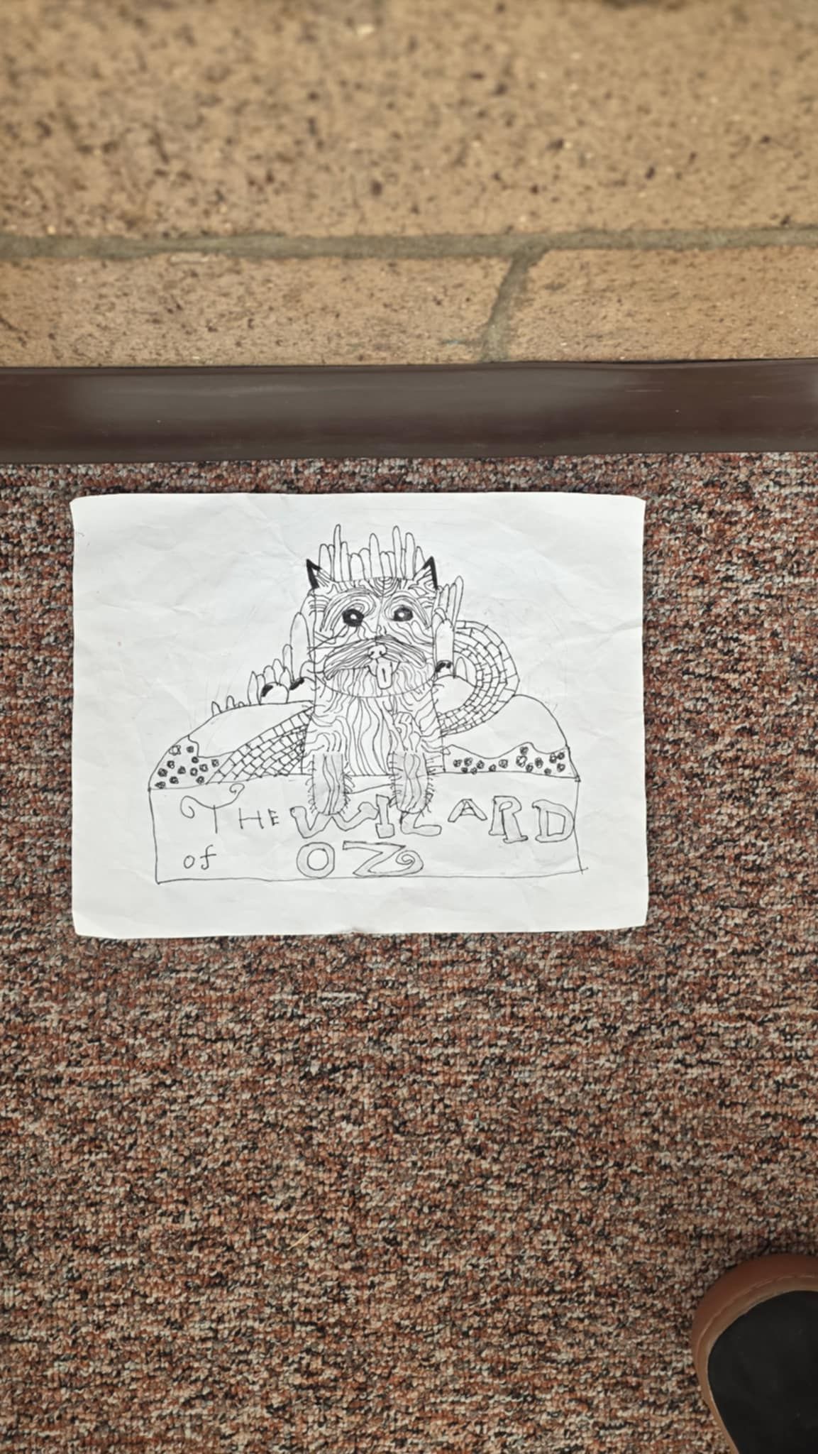 Black and white drawing of a person with a crown sitting on a throne. The image is on a white paper on a brown carpet.