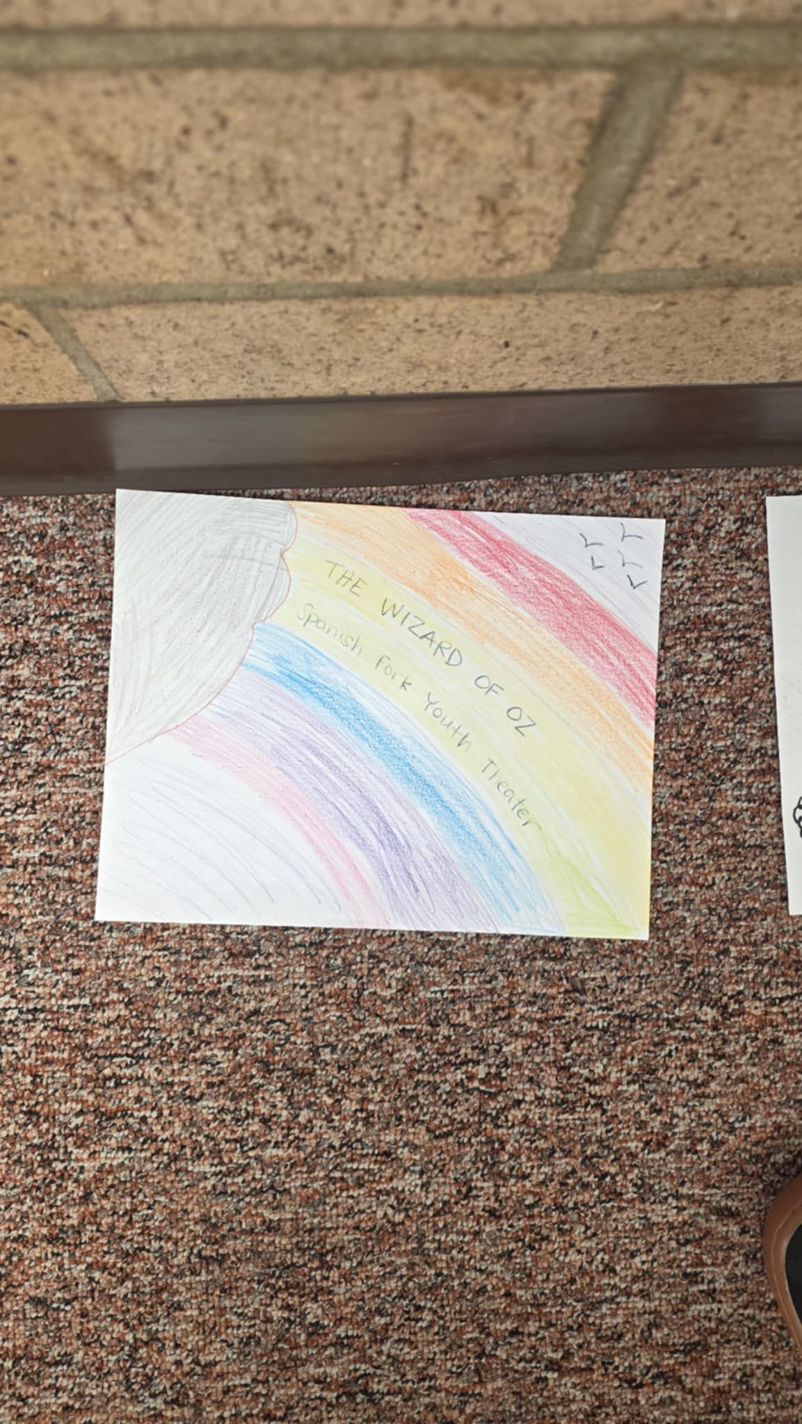 Rainbow drawing on white paper, with handwritten text in the middle, resting on a textured brown surface.