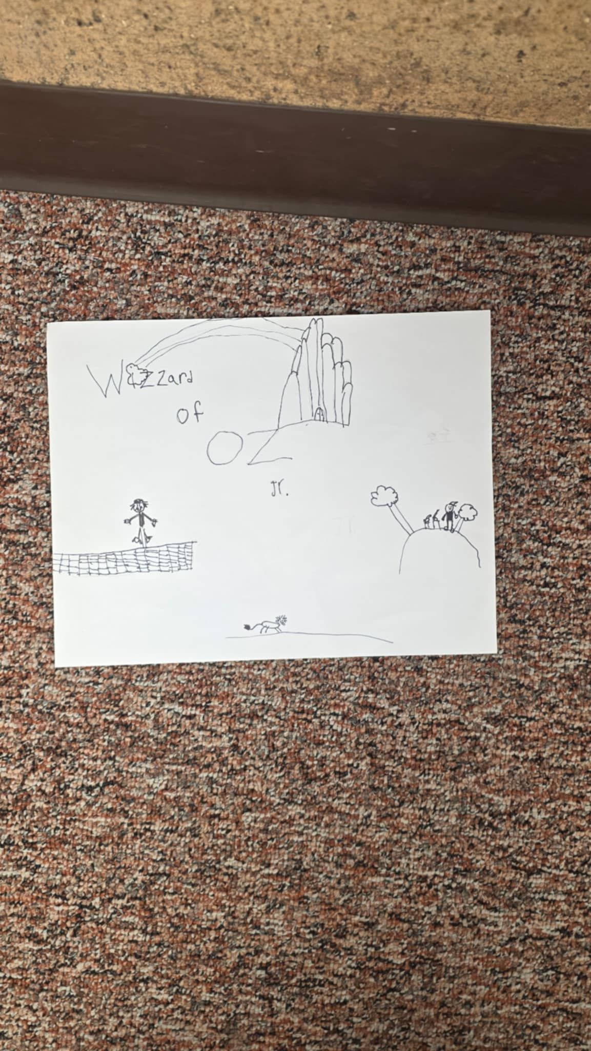 Sketch on paper: a figure stands before buildings, trees, and small shapes are scattered on a brown carpet.