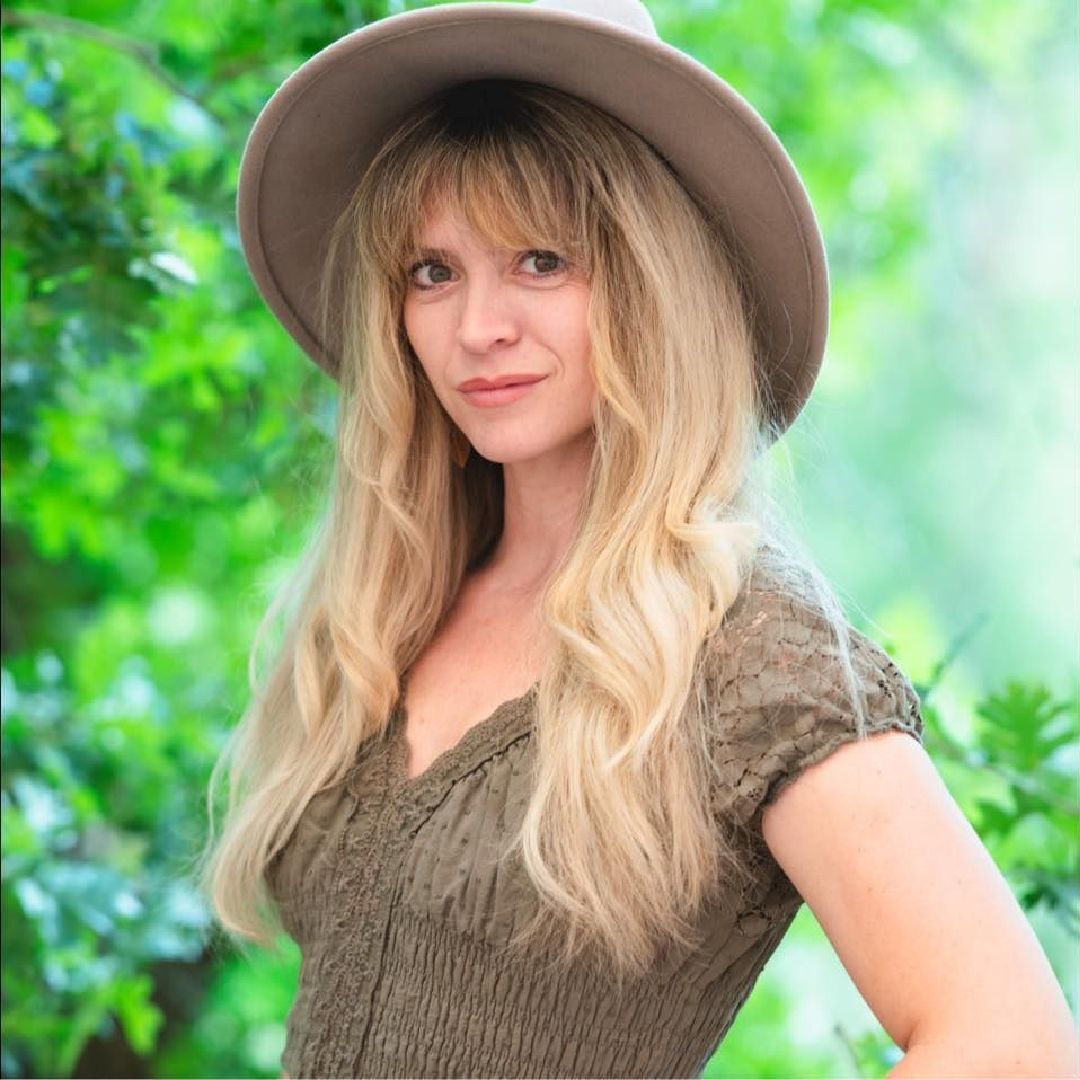A woman wearing a hat and a green dress is standing in front of trees.
