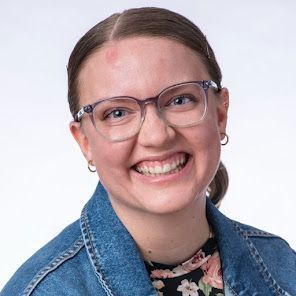 A woman wearing glasses and a denim jacket is smiling.