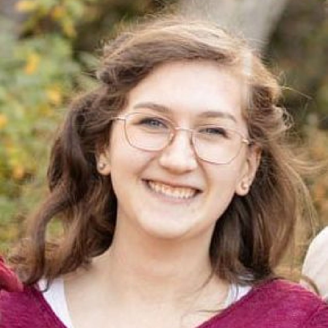 A young woman wearing glasses and a purple sweater is smiling for the camera.