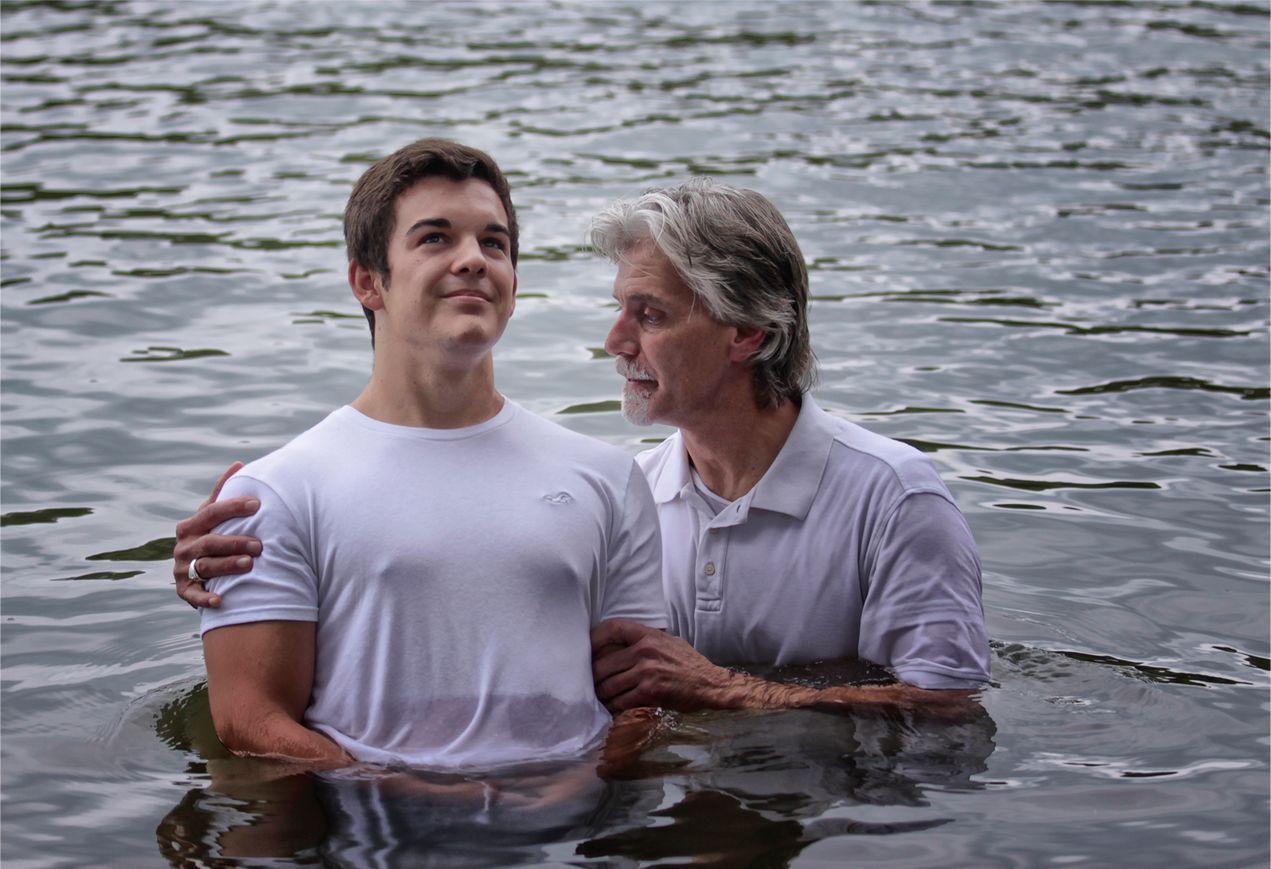 A person is being baptized by another in a calm body of water; both wear white shirts and have serene expressions.