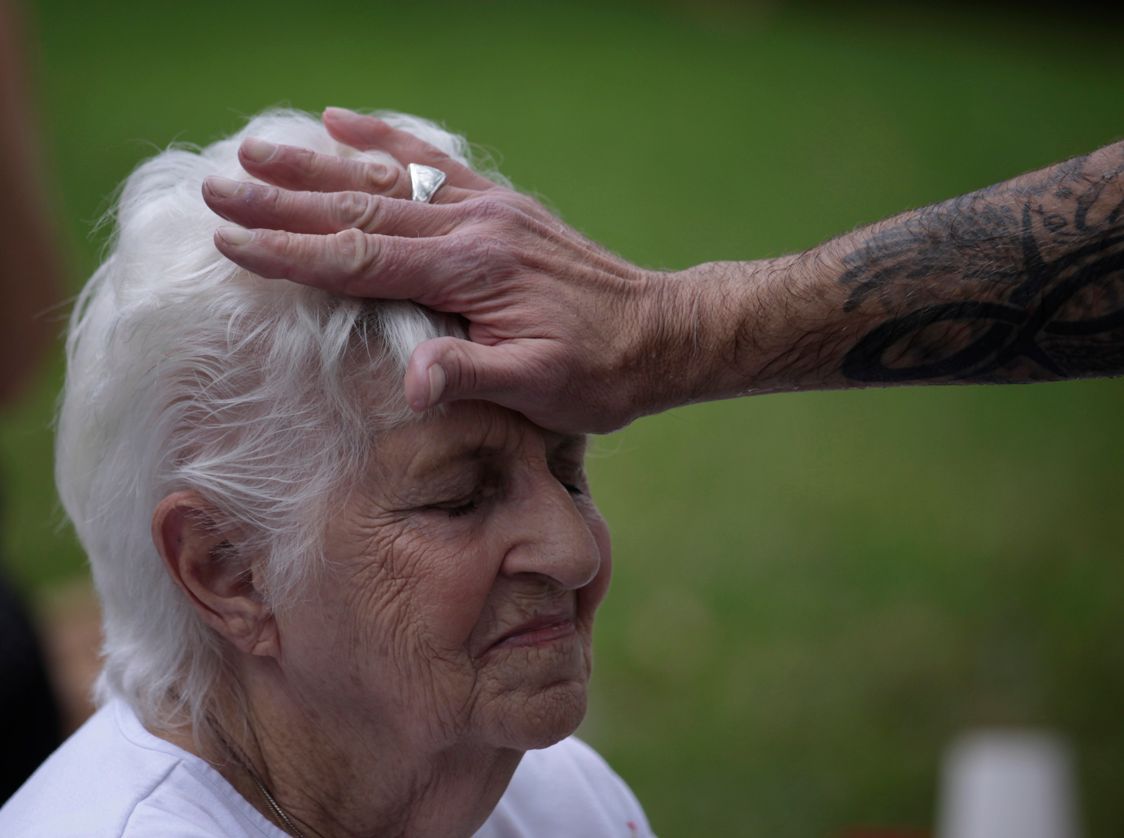 A tattooed hand rests gently on the forehead of a person with closed eyes, outdoors against a blurred green background.