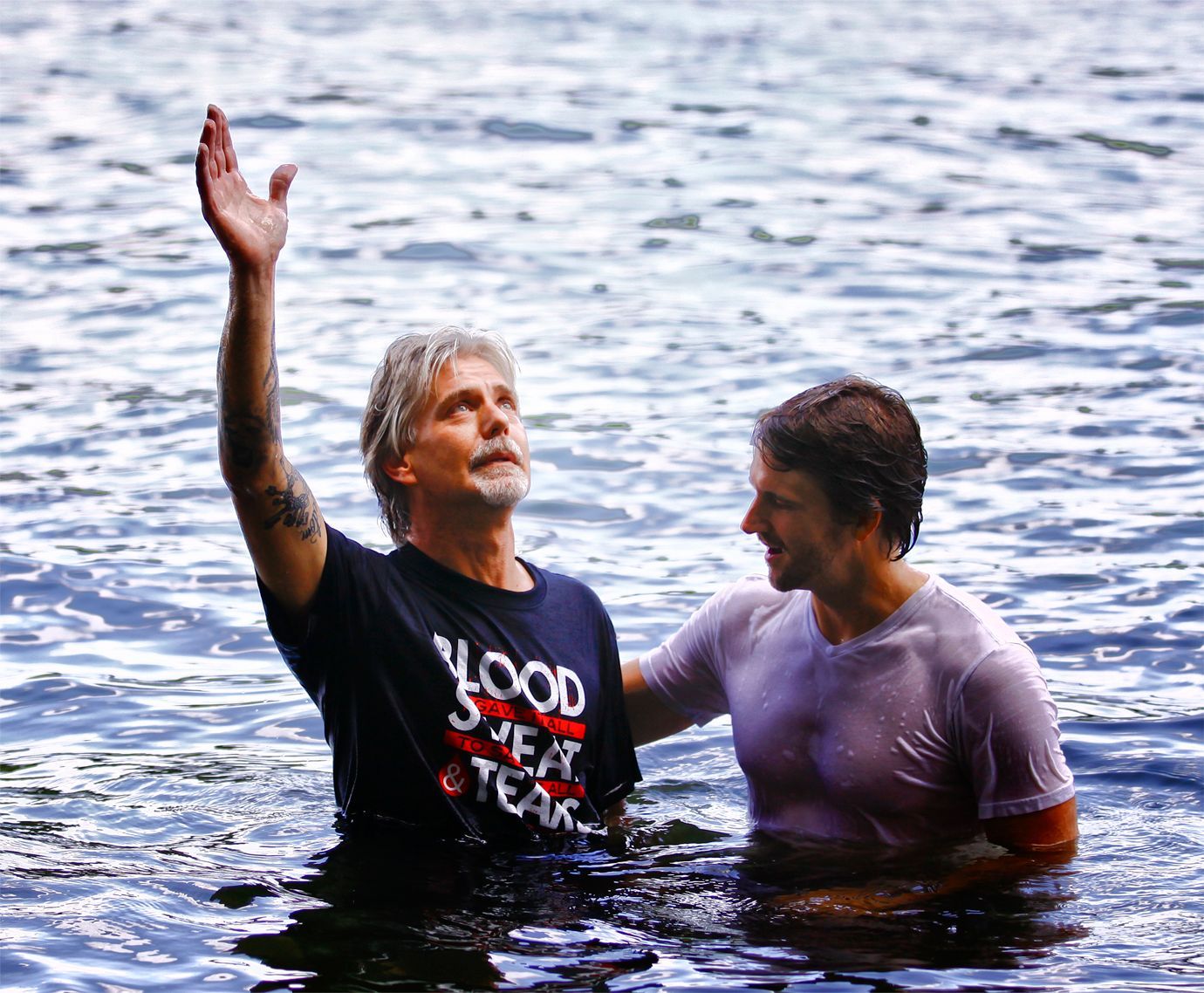Two men standing in a body of water; one with an arm raised upward, the other looking on during a baptism ceremony.