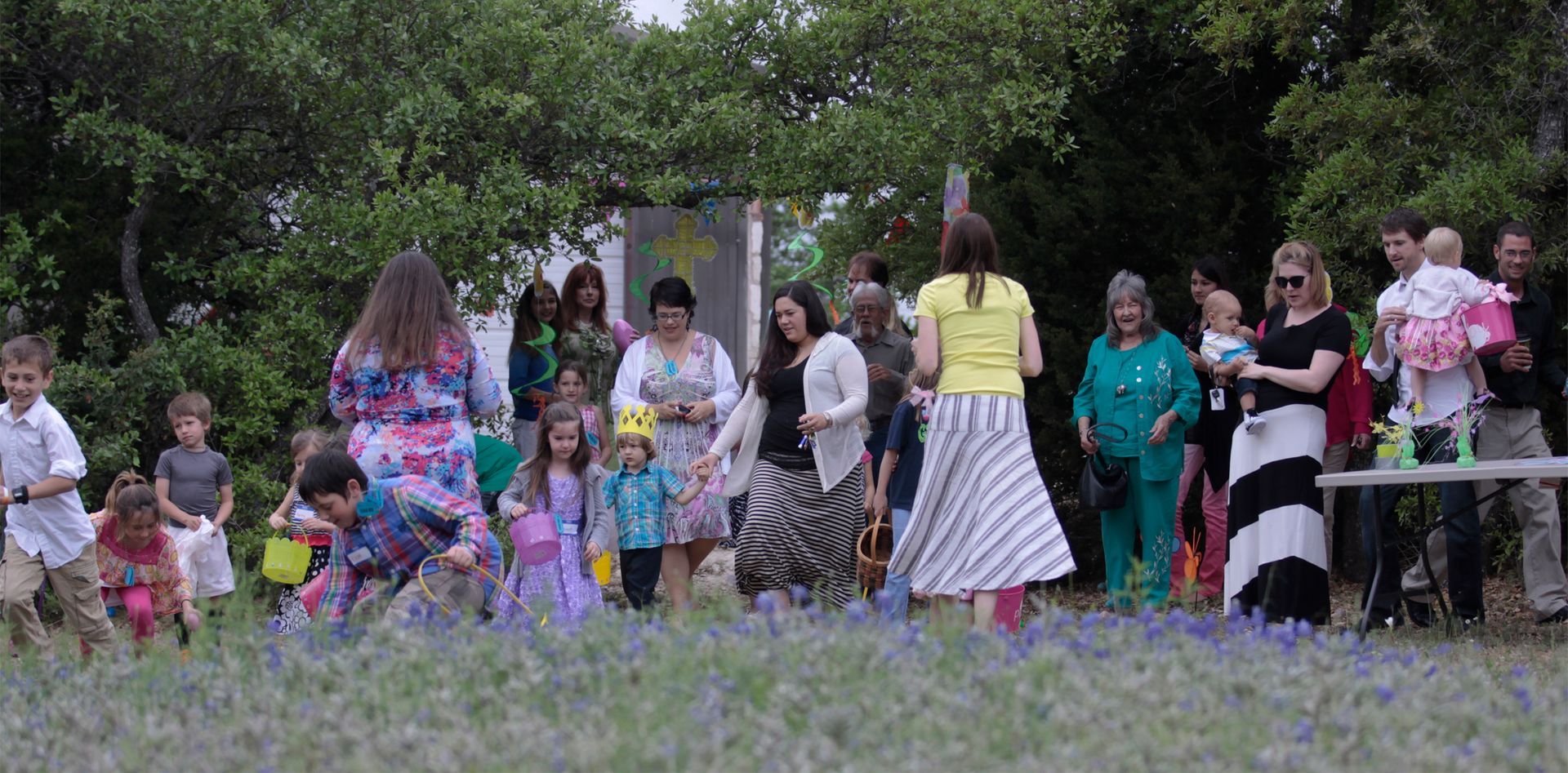 A group of adults and children gather in a park-like, outdoor setting with trees in the background during the day.