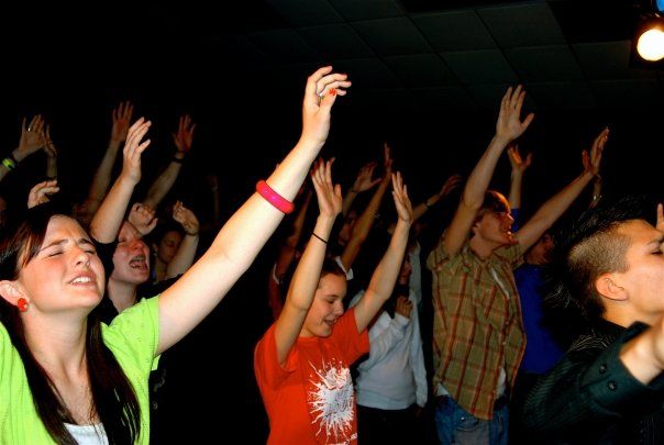 A crowd in a dark space raising their arms in celebration, wearing colorful casual clothing.