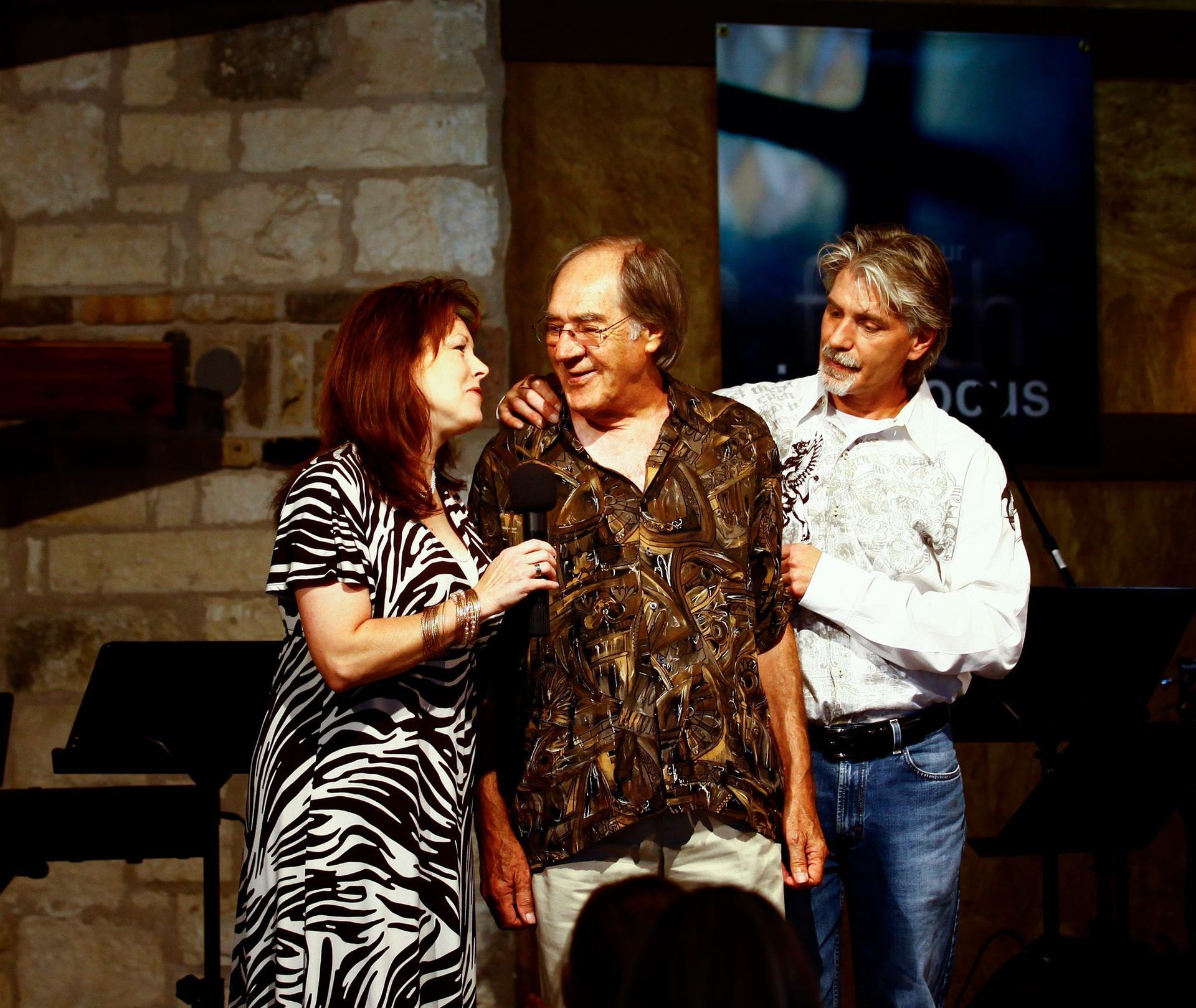 A woman in a zebra-print dress interviews a man on a stage, while another man stands beside him, resting a hand on his back.