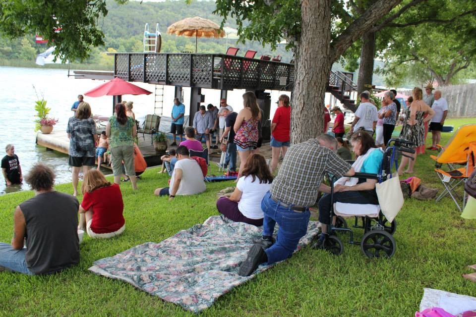 People gather on a grassy lakeside lawn, with some seated on blankets near a wooden deck and others standing nearby.