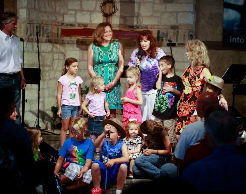 A group of children and adults standing on a stage with stone walls and a wooden cross in a church setting.