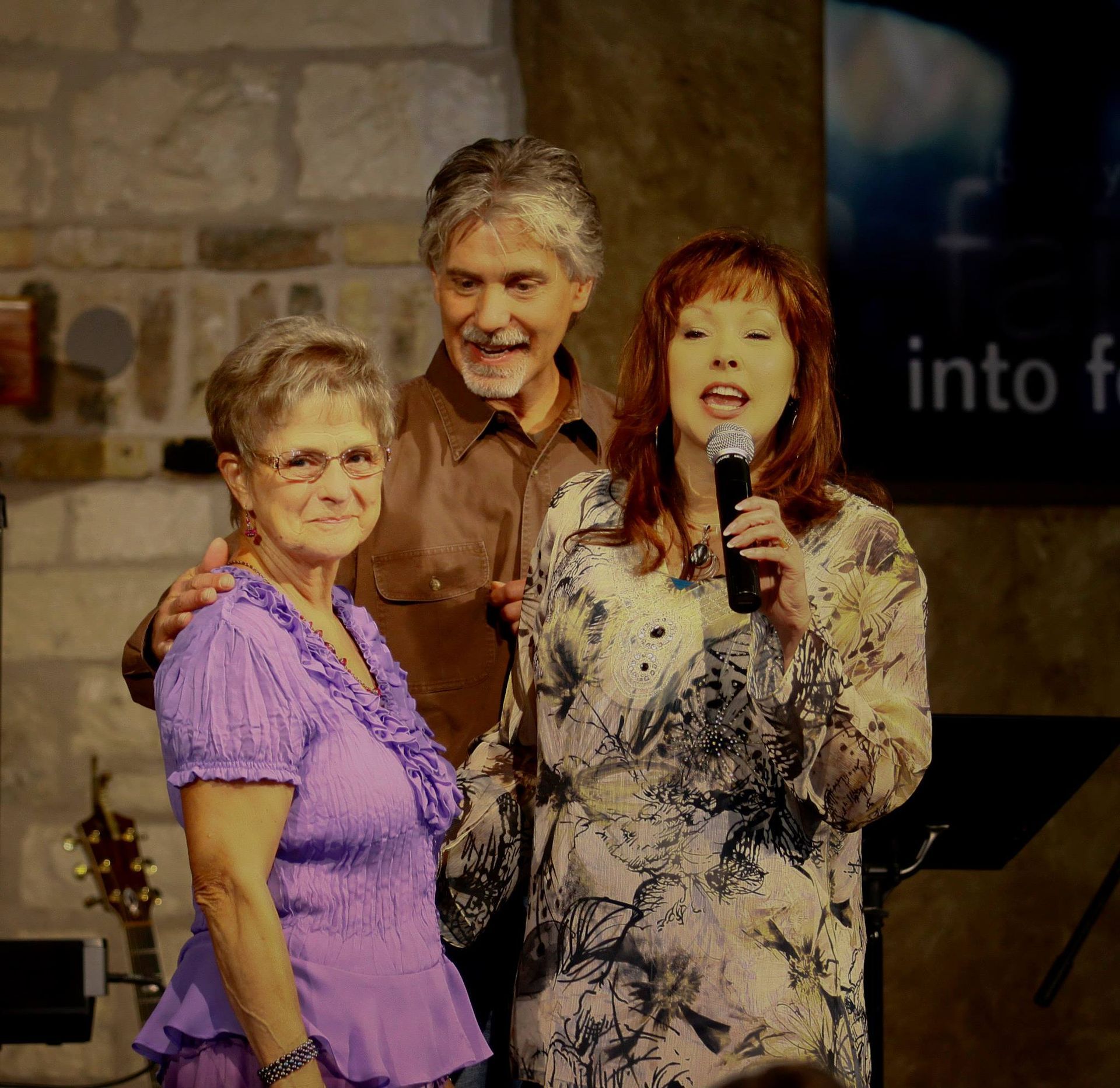 Two people stand on a stage, one singing into a microphone, with a person in a purple top standing in front of them.