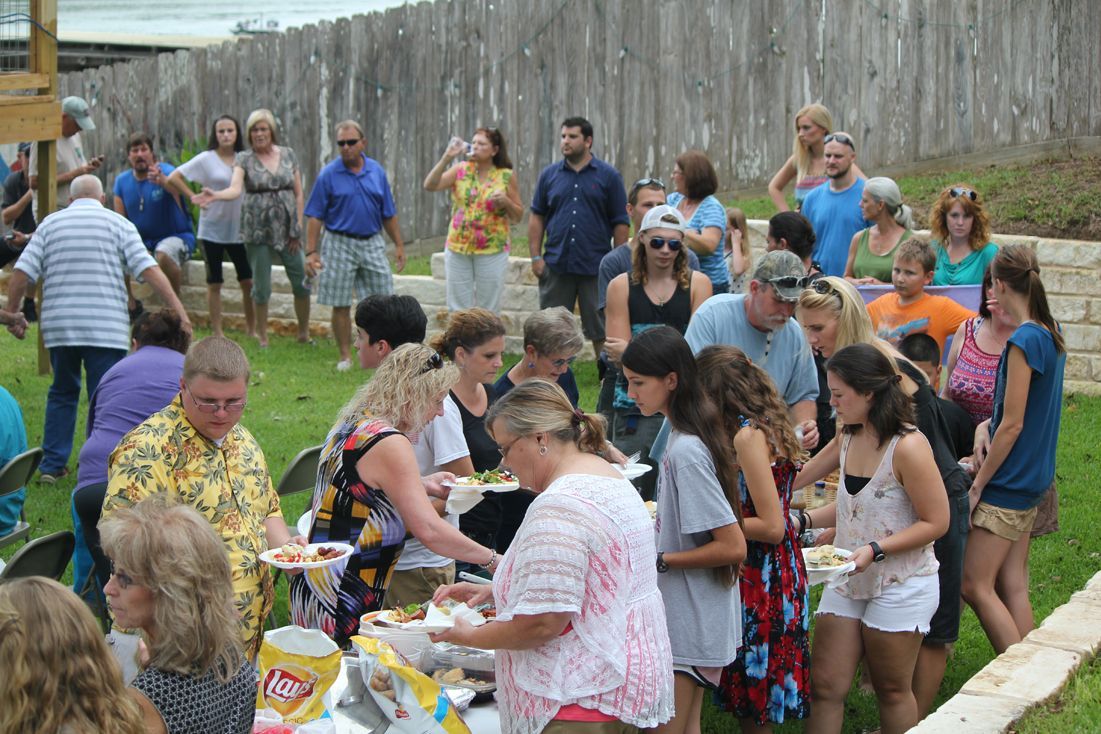 A large group of people gather in a backyard for a buffet-style outdoor meal near a wooden fence.