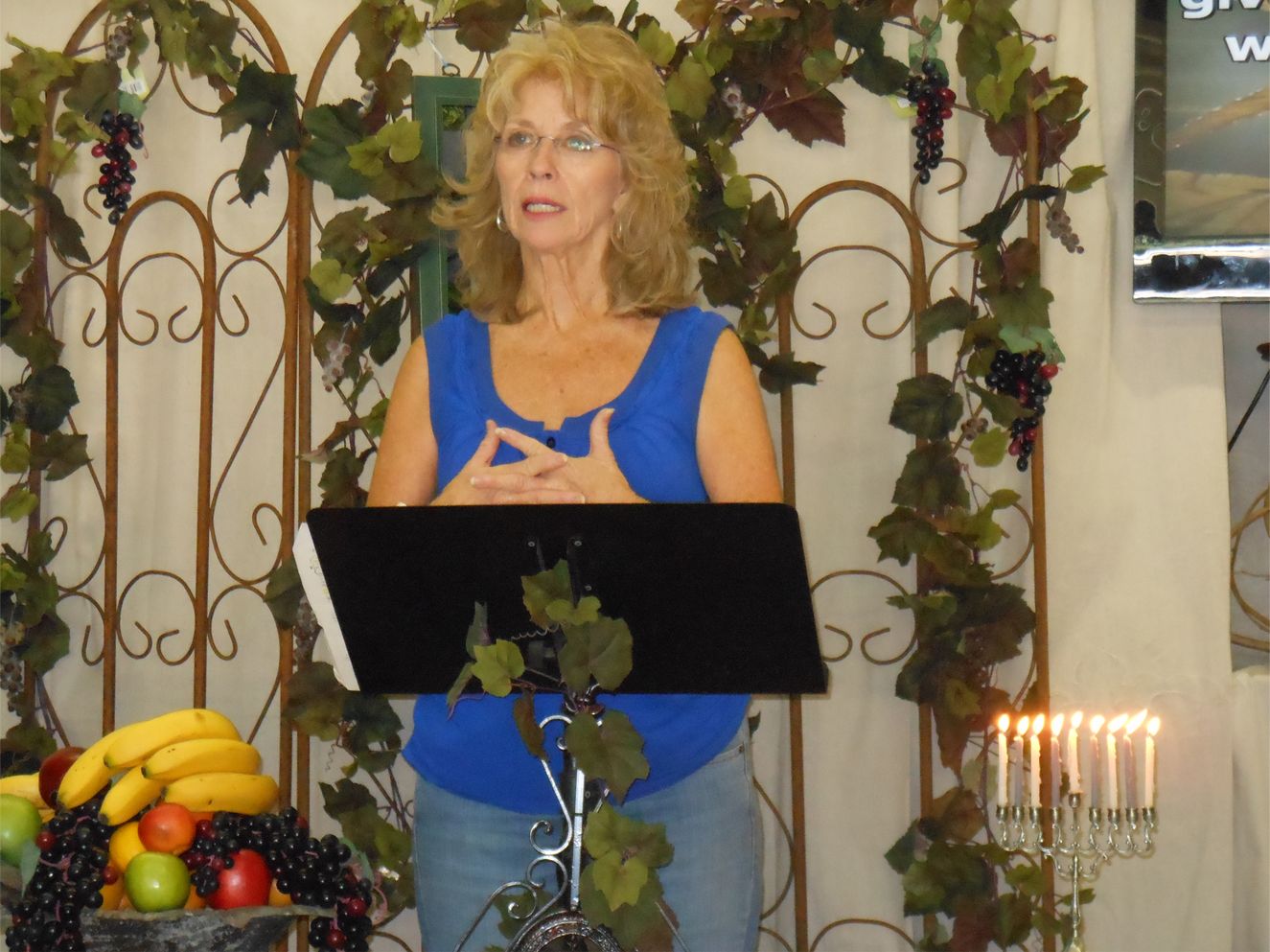 A person stands behind a lectern in front of a decorative trellis with grapes and a lit menorah nearby.