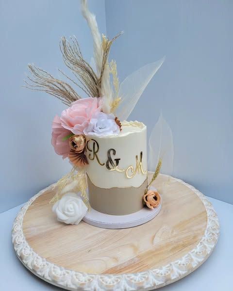 Small wedding cake decorated with flowers, initials, and feathers on a wooden platter.