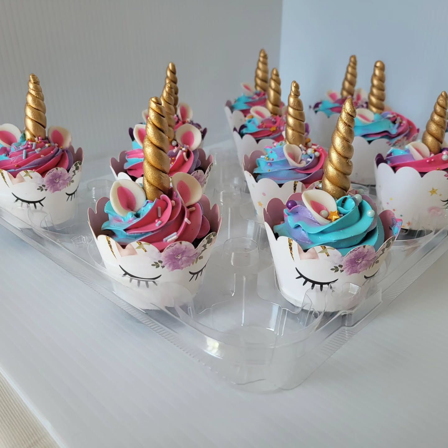 Unicorn cupcakes in a clear plastic container, featuring gold horns, pink and blue frosting, and decorative wrappers.