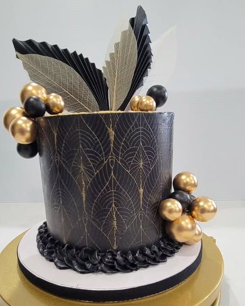 Elegant black and gold cake with leaf design and spheres on a golden stand.