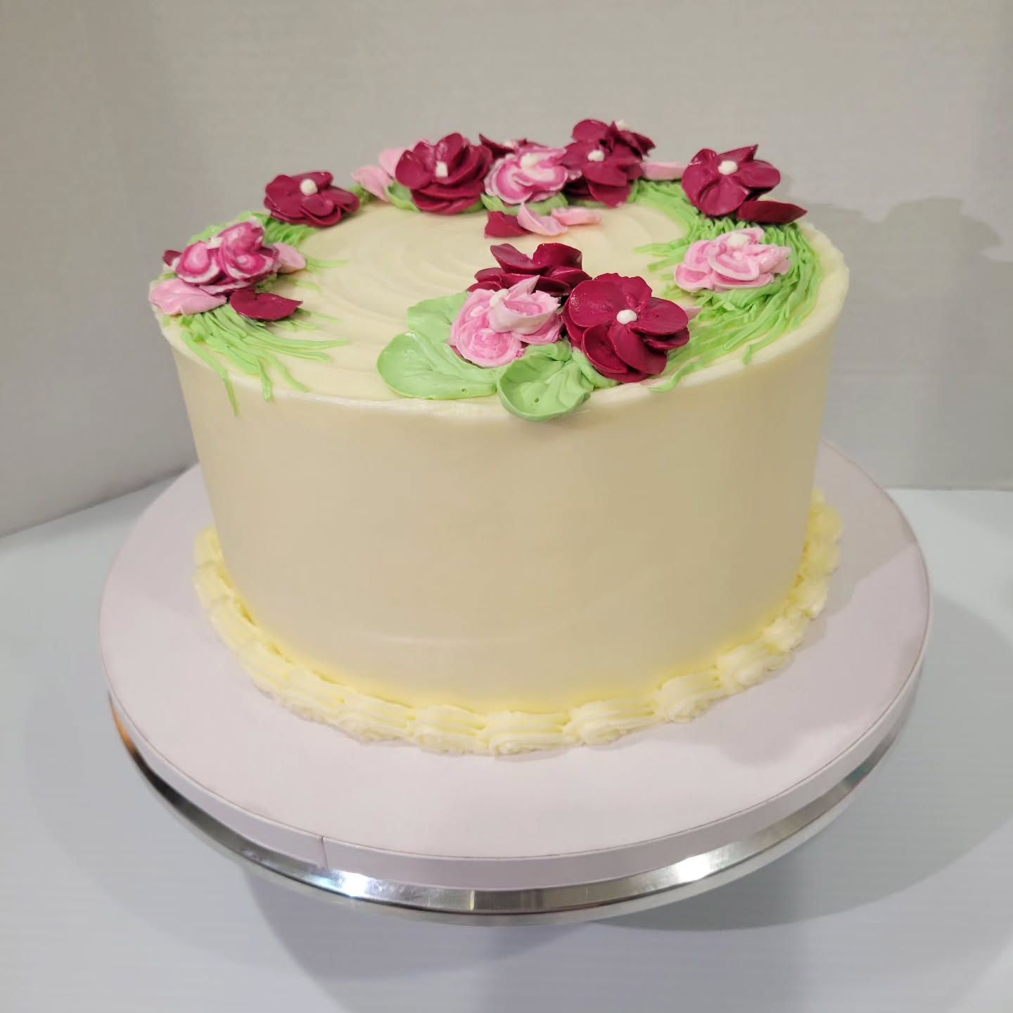 Cake with white frosting, decorated with a floral wreath of pink and burgundy flowers on a light pink cake stand.