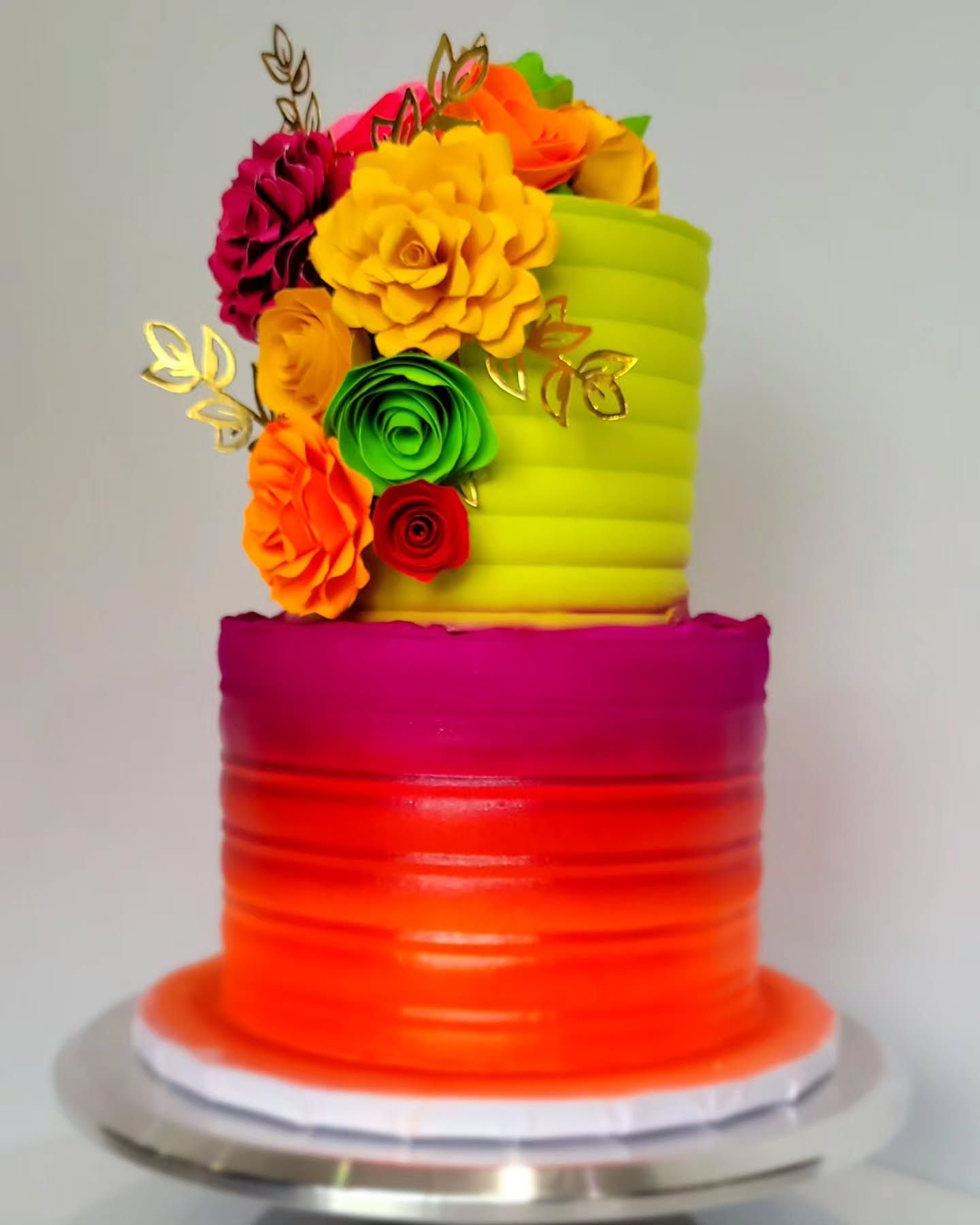 Two-tiered colorful cake with gradient orange-pink bottom and lime green top, decorated with paper flowers and gold accents.