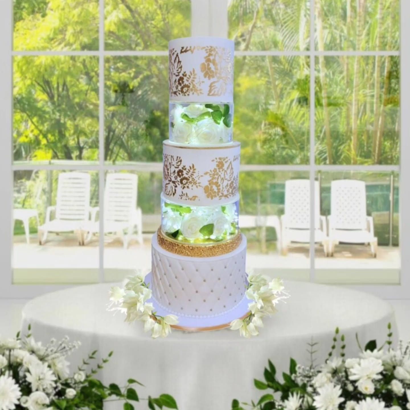 Five-tiered wedding cake on a table, with gold accents, clear layers with flowers, and a window background.