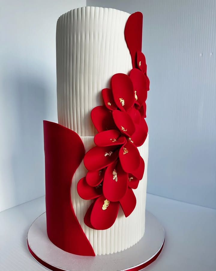 Two-tiered white cake with red accents: vertical ridges, curved side panel, flower petals with gold flecks.