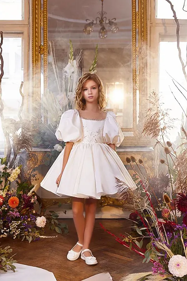 Una bambina vestita di bianco è in piedi davanti a uno specchio circondata da fiori.