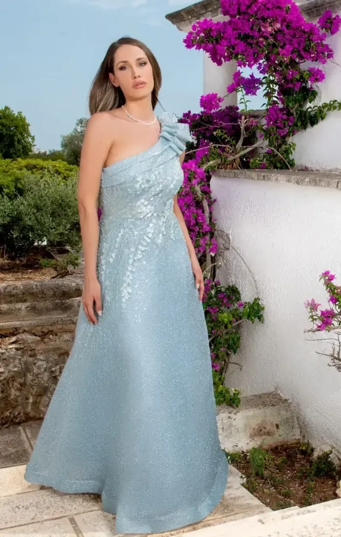 Una donna con un vestito blu monospalla è in piedi davanti a dei fiori viola.